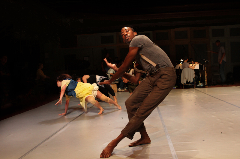 Ricarrdo Valentine wearing brown pants and suspenders dramatically leans his body to one side. There are dancers on the floor in the background.